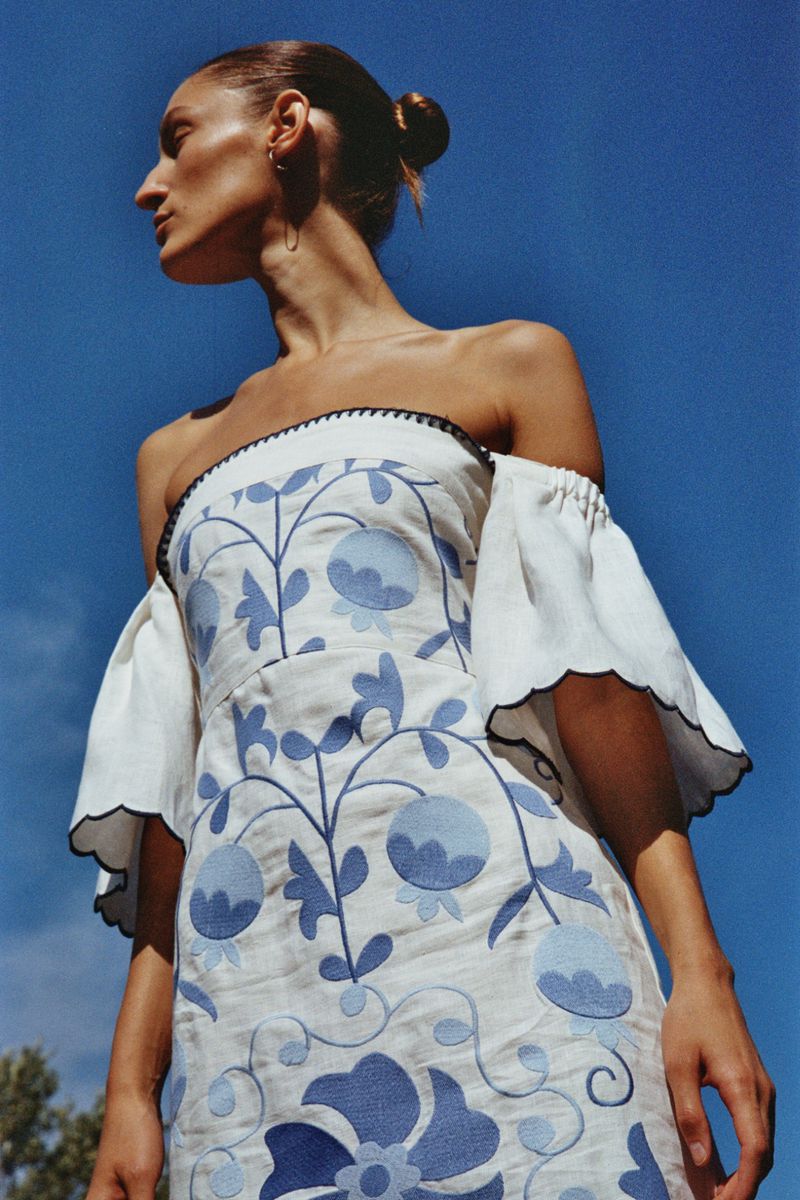 Adult female fashion model poses in a white off-the-shoulder dress with a blue floral pattern against a blue sky backdrop.