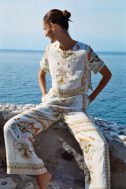 A young woman in a patterned shirt and pants sits on a stone surface against a backdrop of the ocean, smiling and looking away.