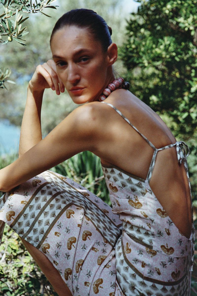 A woman in a patterned dress sits outdoors among green foliage, gazing at the camera with a serene expression, bathed in natural light.