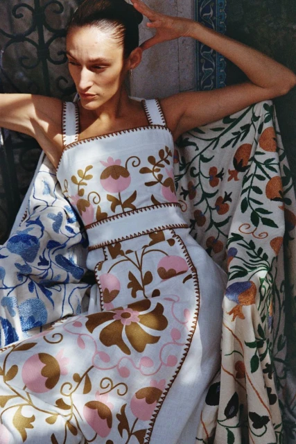 A woman poses in a white dress with floral embroidery, lying on a patterned cloth. Natural light illuminates her and the fabric.