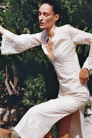 Model Franzi Mueller posing outdoors in an embroidered white lace dress with bell sleeves against lush green foliage.