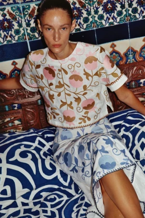 A stylish adult female poses in a floral-embroidered crop top and skirt against a decorative blue-tiled background, seated indoors.