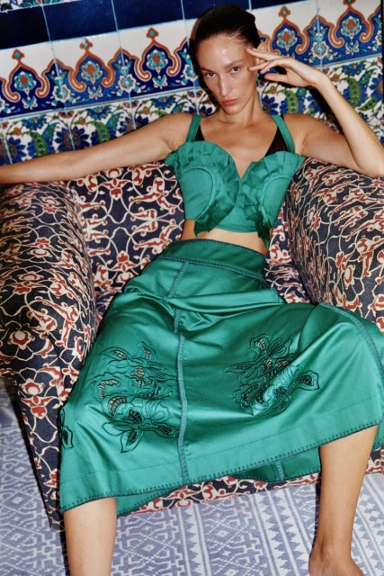 A woman in a green top and skirt sits in a patterned chair against a blue and white tiled wall, with her hand near her face.