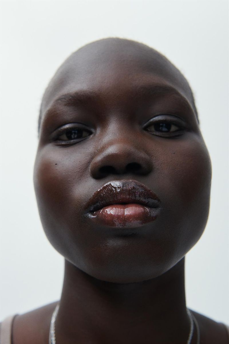 Close-up studio portrait of a dark-skinned woman with shiny lips, minimalist makeup, and a silver chain necklace against a white backdrop.