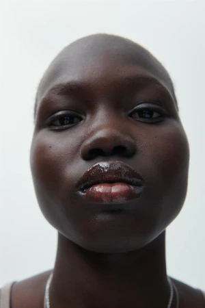 Close-up studio portrait of a dark-skinned woman with shiny lips, minimalist makeup, and a silver chain necklace against a white backdrop.