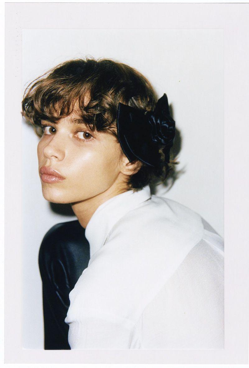 Close-up portrait of a young adult woman with short curly brown hair and a black flower hair accessory, wearing a white shirt.