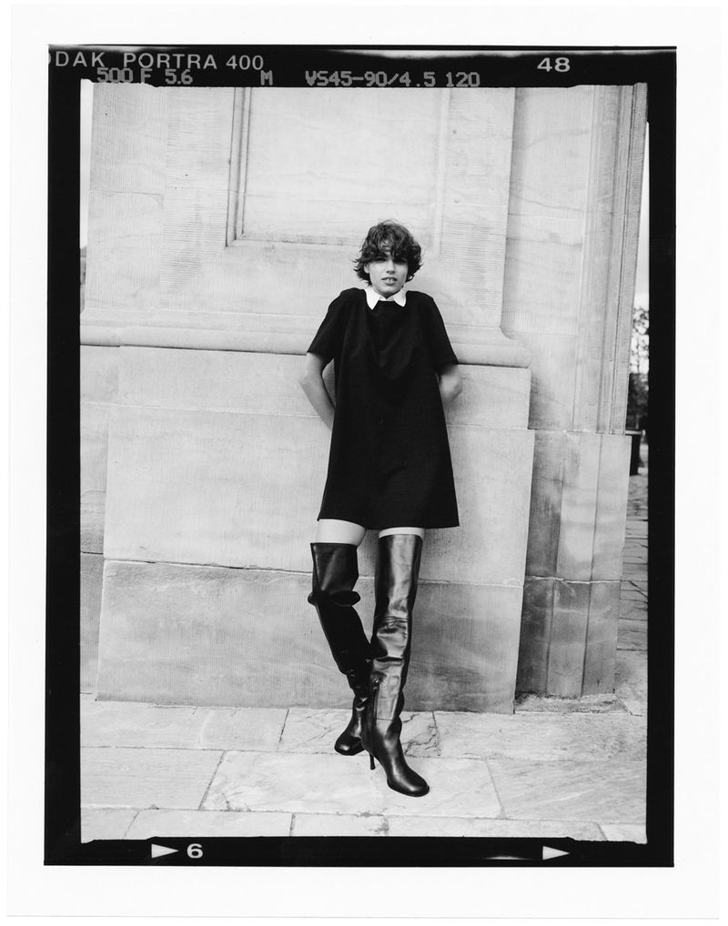 An adult female model poses in a black mini dress and thigh-high leather boots in front of a stone column, captured in black and white.