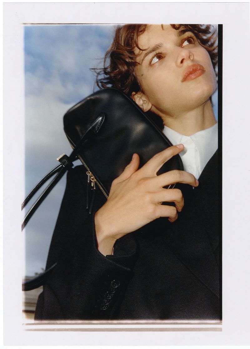 Woman in a black suit and white shirt holds a black leather handbag in front of a cloudy sky for a fashion editorial.