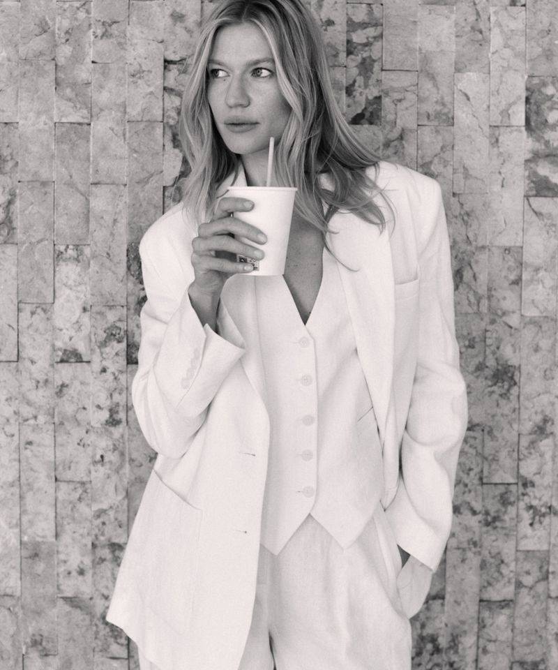 A black and white portrait of a woman in a suit holding a cup with a straw, leaning against a textured wall, with shallow depth of field.