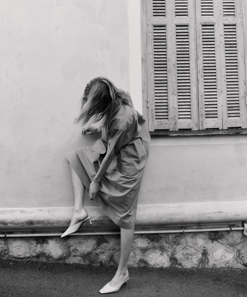 Monochrome fashion shot of a blonde woman posing with her leg up against a wall while wearing a dress and heels in an urban setting.