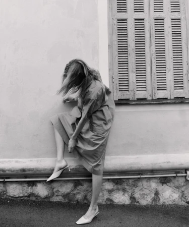 Monochrome fashion shot of a blonde woman posing with her leg up against a wall while wearing a dress and heels in an urban setting.