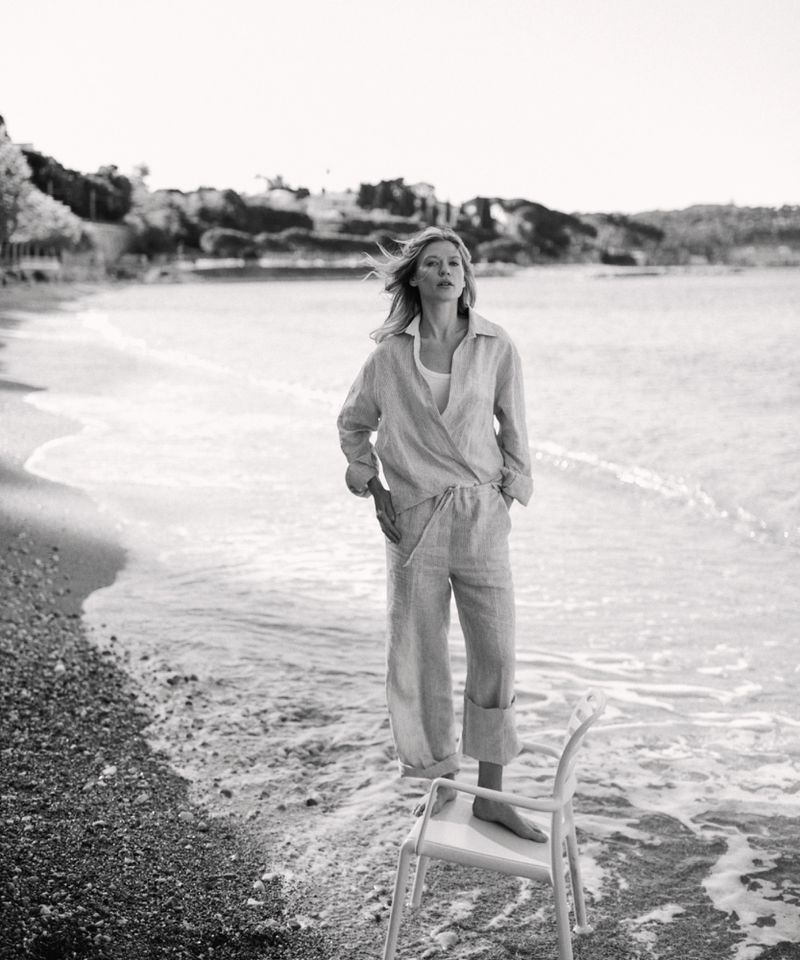 Monochromatic shot of woman in light-colored linen set on beach, standing on white chair near water, ocean, shoreline, and sandy beach.