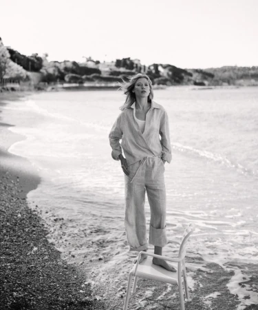 Monochromatic shot of woman in light-colored linen set on beach, standing on white chair near water, ocean, shoreline, and sandy beach.