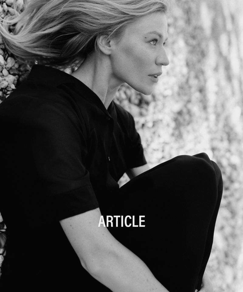 Black and white portrait of an adult woman with blonde hair wearing a dark top, posing outdoors against a pebbled background.