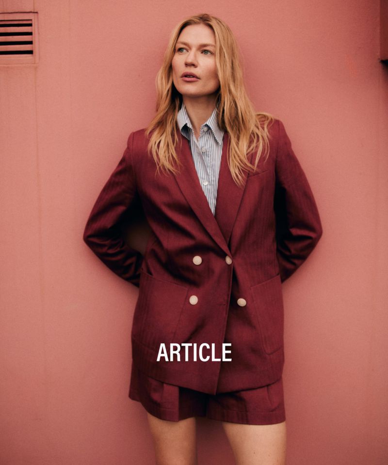 A fashionable blonde woman poses in a brick pink photo studio wearing a burgundy skirt suit with a light blue striped button-down shirt.