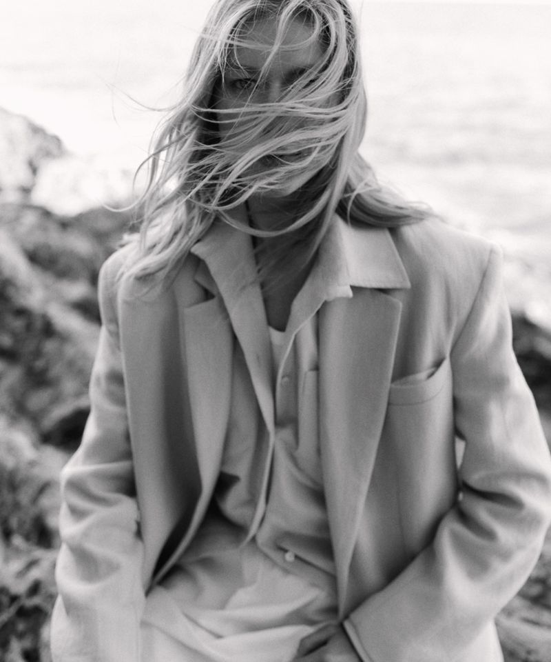 Monochrome portrait of a fair-skinned woman with blonde hair blowing in the wind, wearing a blazer on a rocky coast near the ocean.