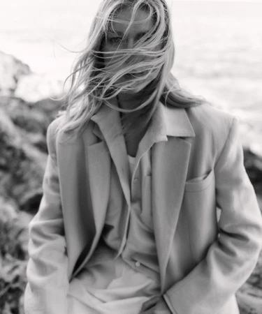 Monochrome portrait of a fair-skinned woman with blonde hair blowing in the wind, wearing a blazer on a rocky coast near the ocean.