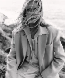 Monochrome portrait of a woman with long hair blowing across her face wearing a tailored blazer outdoors by the water.