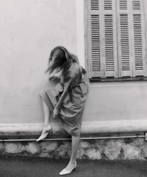 Woman in dress leaning against a plastered building wall beneath wooden shutters adjusting her heeled shoe