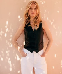 Blonde woman wearing a black vest and white pants posing against a textured wall with dappled sunlight shadows