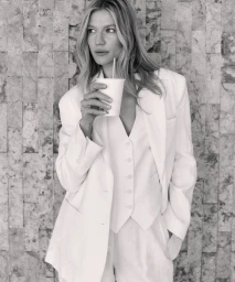 Black and white portrait of a blonde woman in a white suit holding a paper cup against a stone wall.