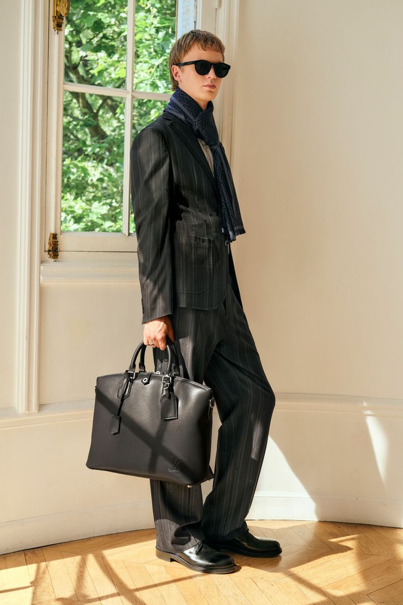 Full-length shot of a stylish adult male wearing a pinstripe suit, scarf, sunglasses, and leather shoes, holding a briefcase indoors.