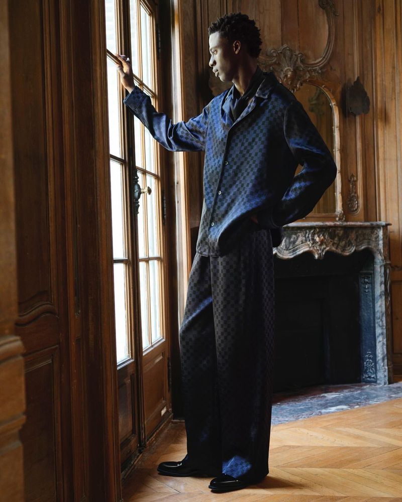 An adult male in a patterned lounge set stands inside, gazing out of a large window in a traditionally styled room with wood panelling.
