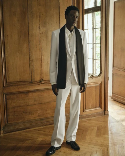 A slender, dark-skinned man poses in a stylish white suit with a black scarf draped around his neck, set against an ornate wood background.