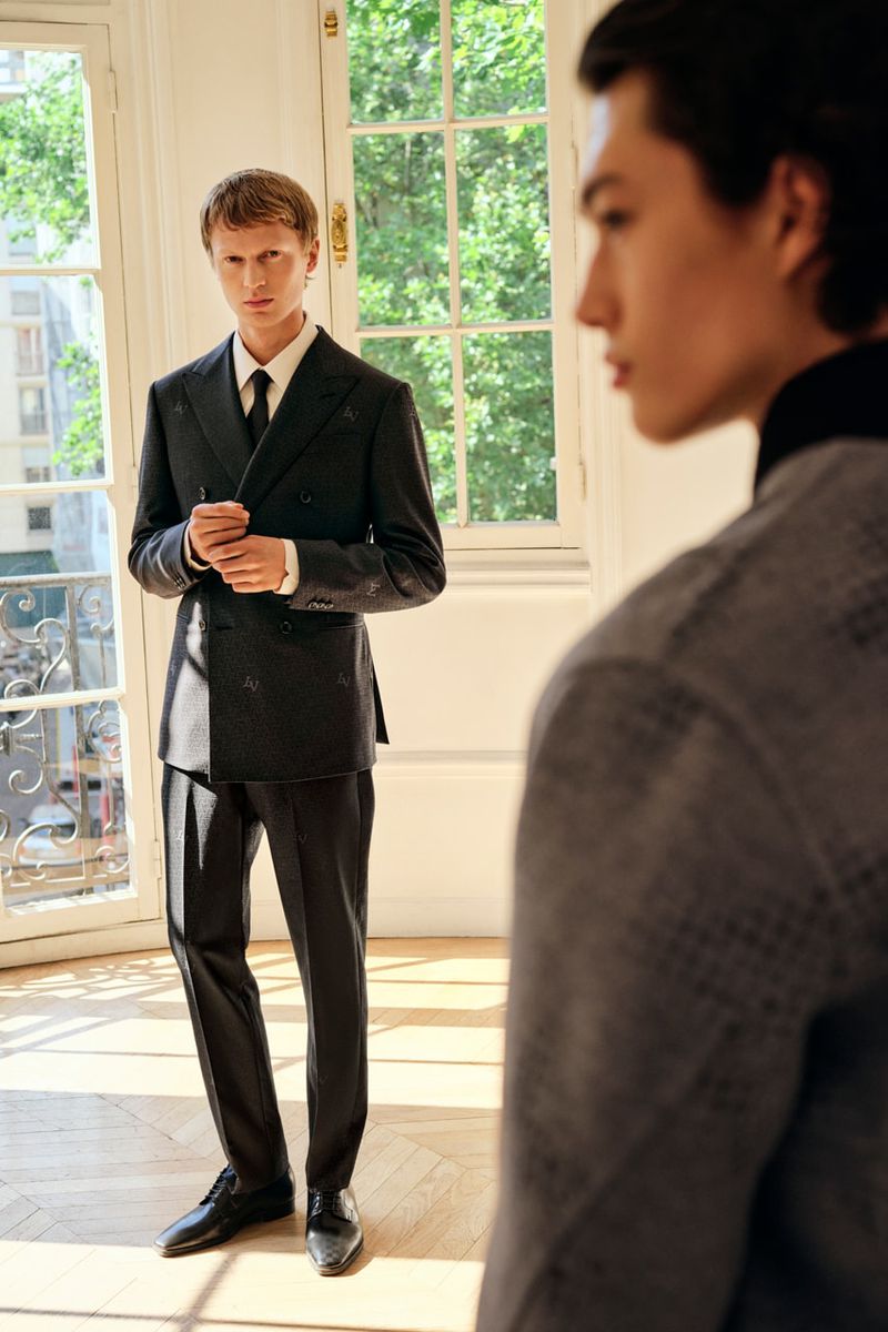Two men are indoors with a window view; one is in focus, in a suit, while the other, partially visible, wears a grey jacket.