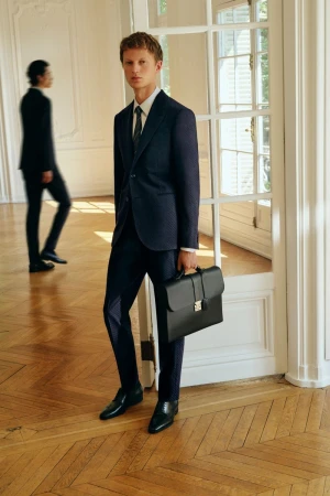 Elegant male models in suits pose indoors near windows. One holds a briefcase on a parquet floor in a light-filled setting.