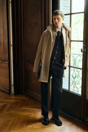 Man in a tan trench coat and black suit stands inside by a window. Dark wood floors add to the indoor fashion photograph.