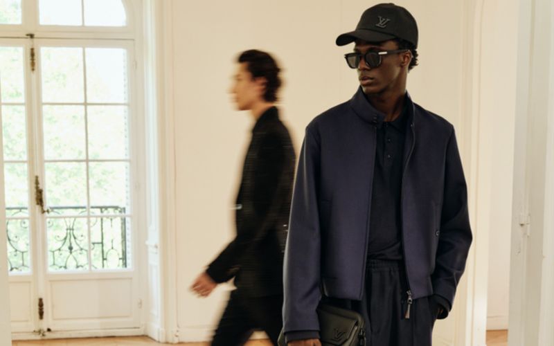 Fashion shot of an adult male model wearing a blue jacket, black cap, and sunglasses, with another man blurred in the background.