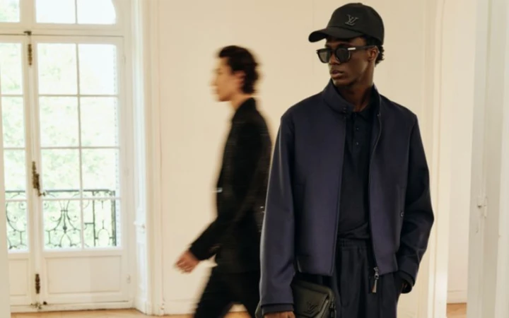 Fashion shot of an adult male model wearing a blue jacket, black cap, and sunglasses, with another man blurred in the background.