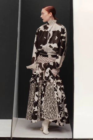 A red-haired fashion model poses in a studio with black and white partitions, wearing a brown and white cow-print top and tiered skirt.