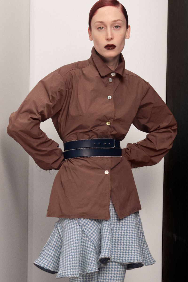 Adult woman with red hair poses in studio wearing brown shirt, wide black belt, and patterned skirt against white and black background.