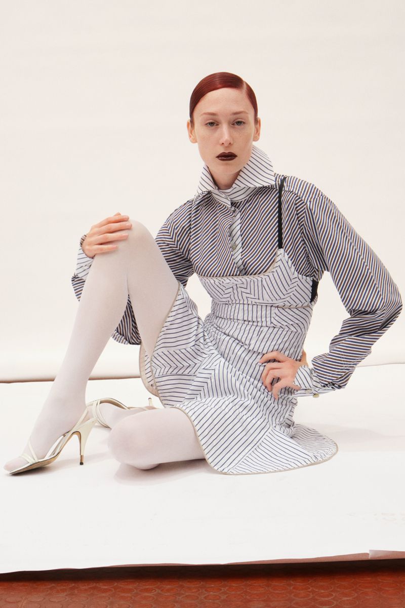 Fashion editorial shot of a red-haired adult woman in a striped dress and shirt, white tights and high heels, sitting on a white surface.
