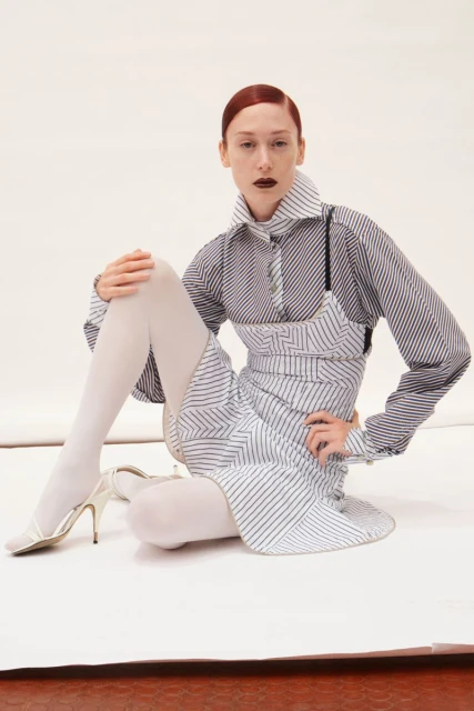 Fashion editorial shot of a red-haired adult woman in a striped dress and shirt, white tights and high heels, sitting on a white surface.