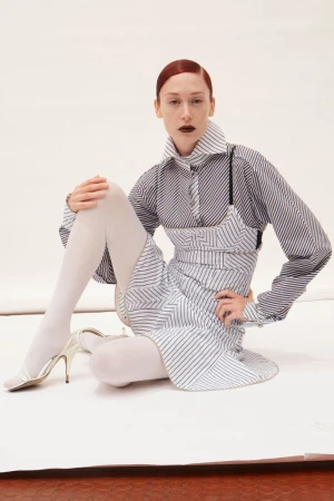 Fashion editorial shot of a red-haired adult woman in a striped dress and shirt, white tights and high heels, sitting on a white surface.