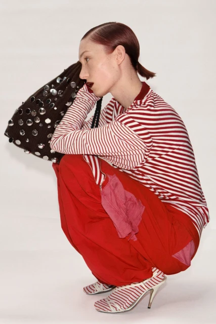 Profile shot of a fair-skinned woman in striped top and red pants, squatting in striped heels, holding a beaded bag in a white studio.