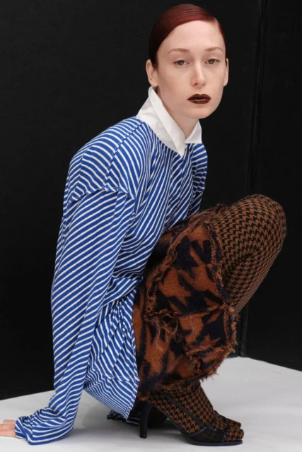 Adult woman in blue striped shirt and houndstooth print leggings squats indoors in front of a black and white background.