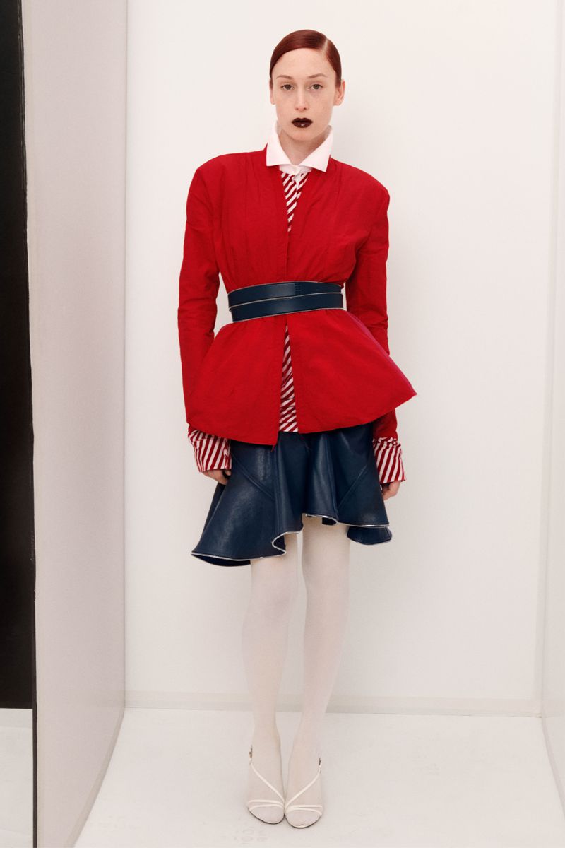 Fashion photo of a woman with red hair and dark lipstick wearing a red jacket, blue skirt, and white heels in a white room.