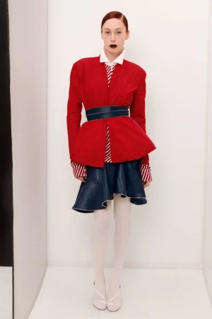 Fashion photo of a woman with red hair and dark lipstick wearing a red jacket, blue skirt, and white heels in a white room.