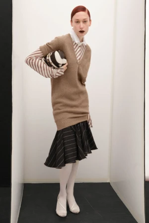 Fashionable adult female poses indoors wearing a beige sweater, white striped skirt and shirt, white tights, and white shoes while holding ball.