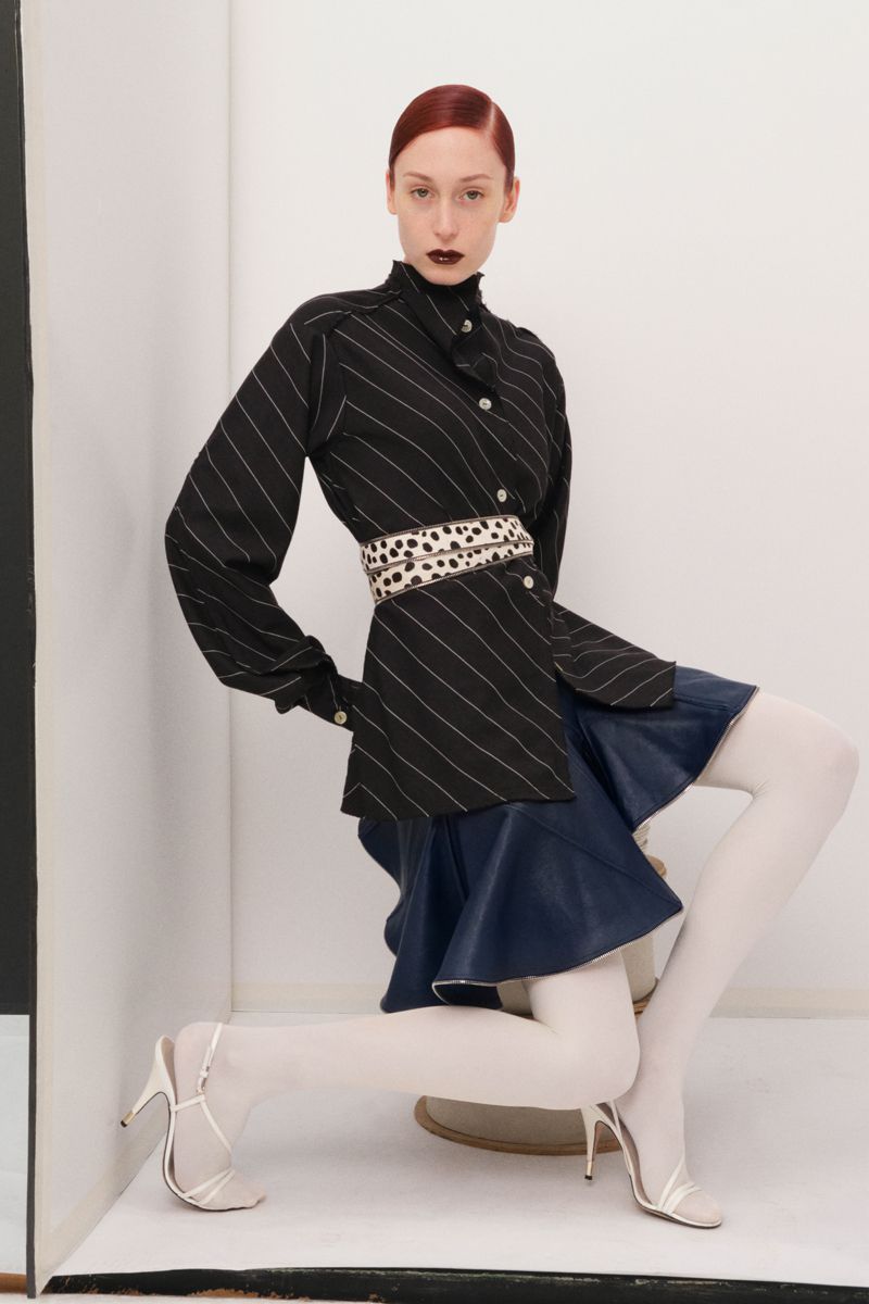 A slender woman with auburn hair strikes a dynamic pose in a striped black blouse, blue skirt, white tights, and matching stilettos in studio.