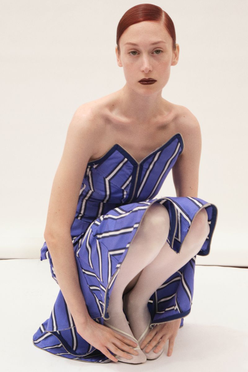 Elegant red-haired woman in a striped blue and white dress crouching indoors against a plain white backdrop in a fashion shot.