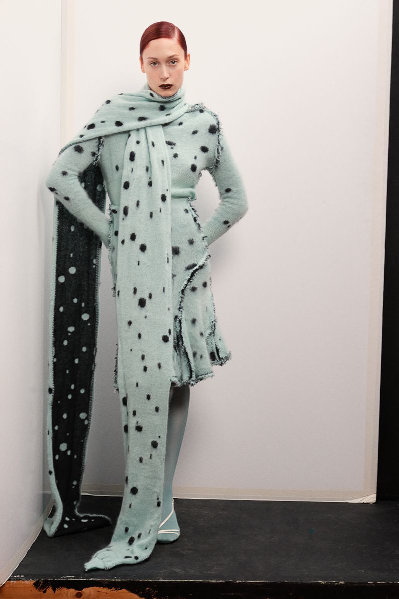 A single woman poses in a studio wearing a light green polka-dotted knit dress and a matching scarf, casting a fashionable pose.