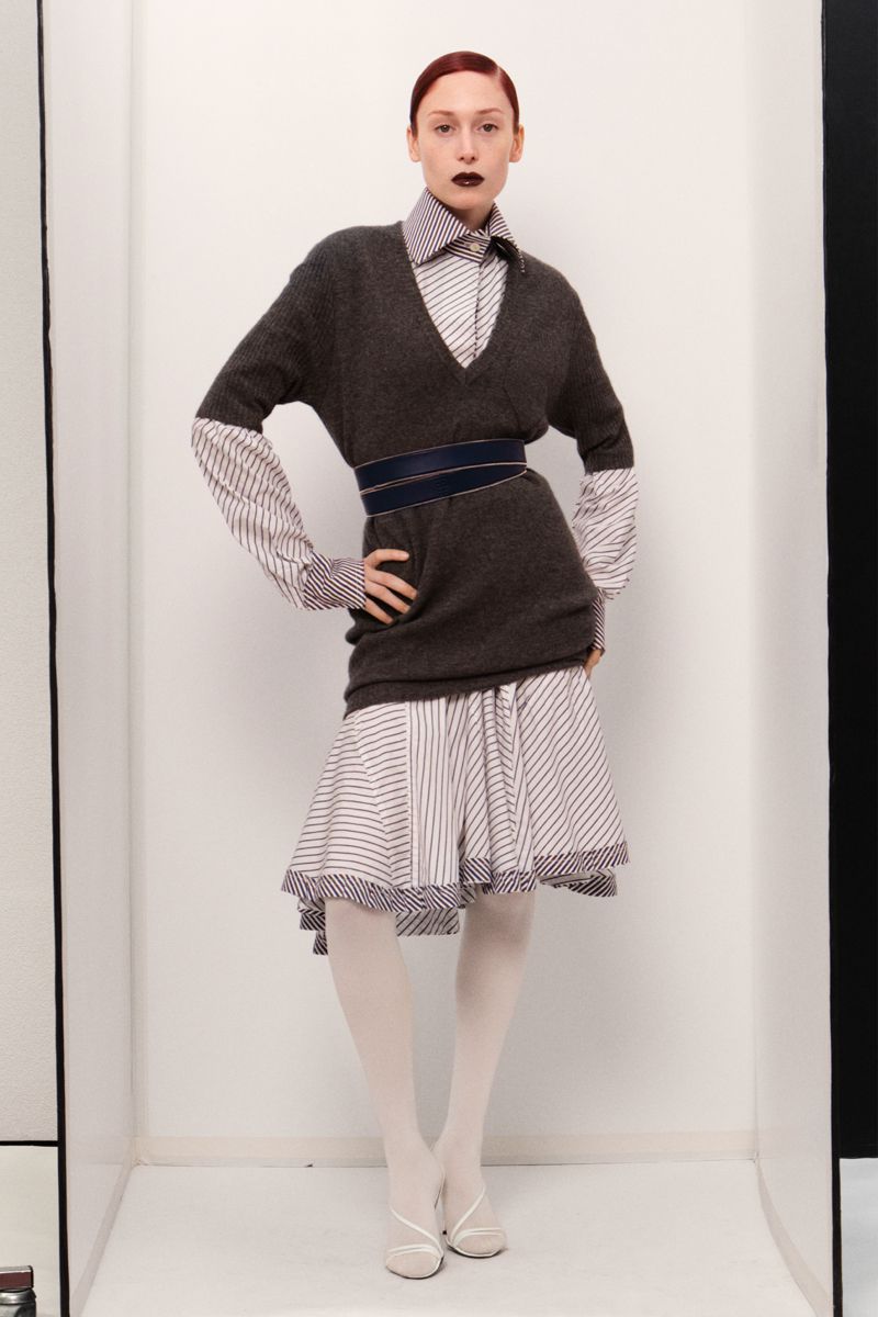 Elegant adult woman poses in a studio wearing a grey sweater, striped skirt, and white tights, hands on her hips, showcasing fashion.