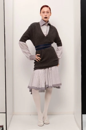 Elegant adult woman poses in a studio wearing a grey sweater, striped skirt, and white tights, hands on her hips, showcasing fashion.