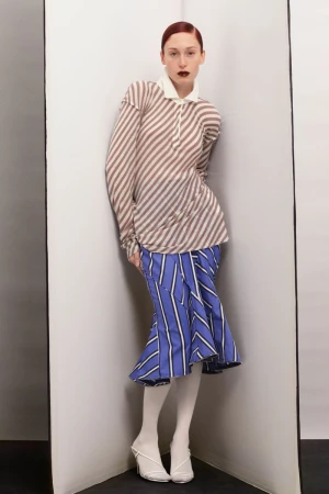 A woman in a striped top and skirt poses in a corner. She wears white tights and heels in a studio setting with neutral colours.