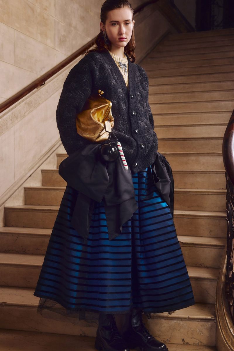 Elegant woman in a black cardigan and blue-striped skirt poses on stairs with a golden clutch, showcasing fashion-forward style.